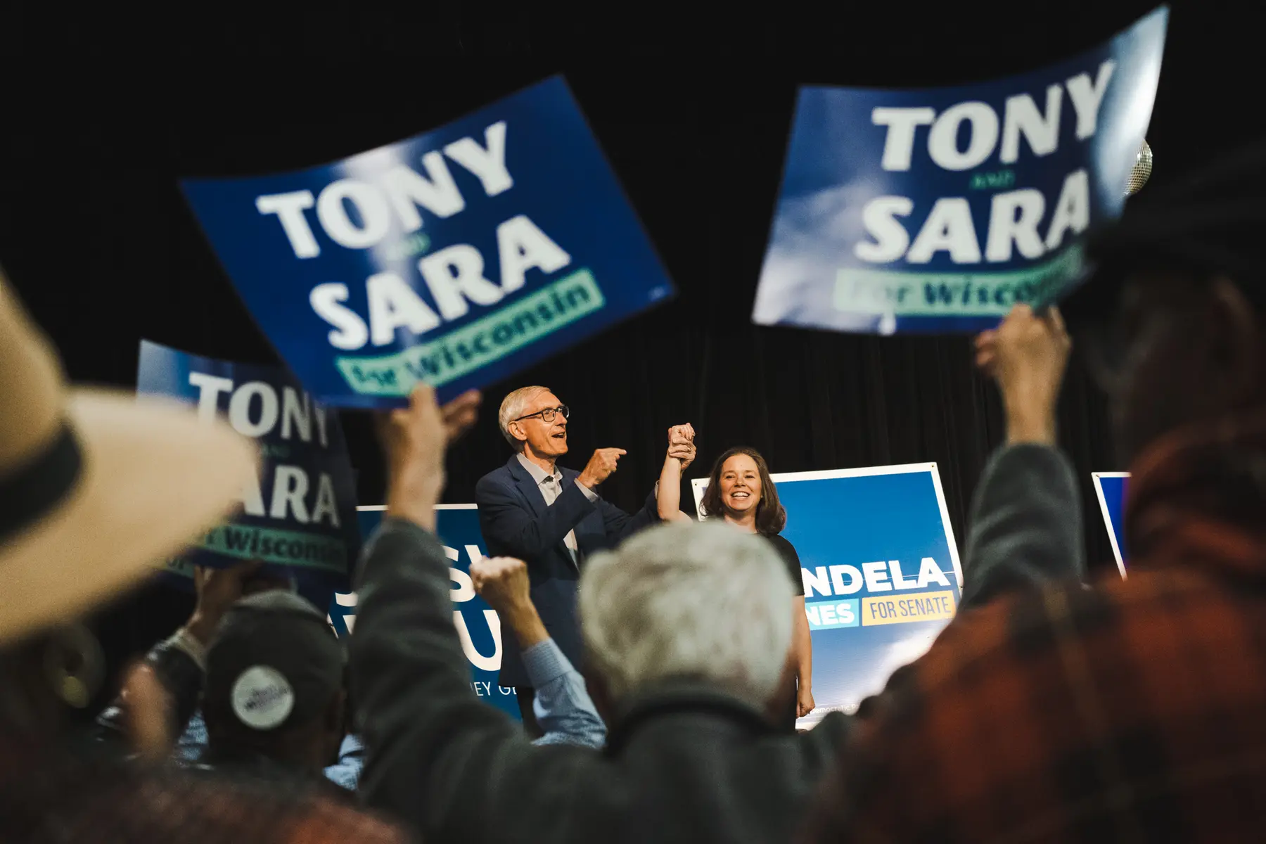 Tony Evers und Sara Rodriguez stehen auf einer Bühne, während Menschen Schilder mit ihren Namen hochhalten.