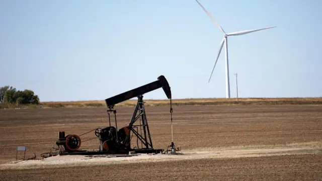 Eine Ölpumpanlage in einer weiten Landschaft mit einer Windkraftanlage im Hintergrund.