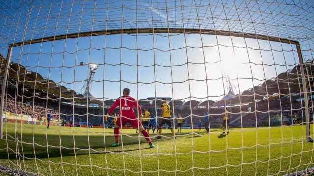 Een foto van de achterkant van een voetbal doel met een goalkeeper en spelers op het veld