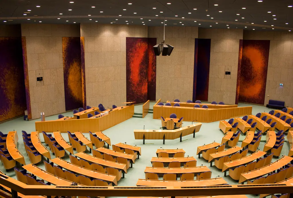 Overzicht van de plenaire zaal van de Tweede Kamer met houten banken en kleurrijke wandkleden.