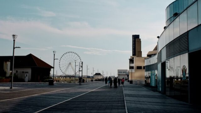 Wide wooden boardwalk with a ferris wheel in the distance and modern glass-fronted buildings along the right side under a clear sky