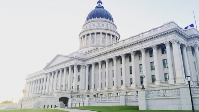 A white stone government building with tall columns, a large domed roof, and a curved walkway across a green lawn under a clear sky.