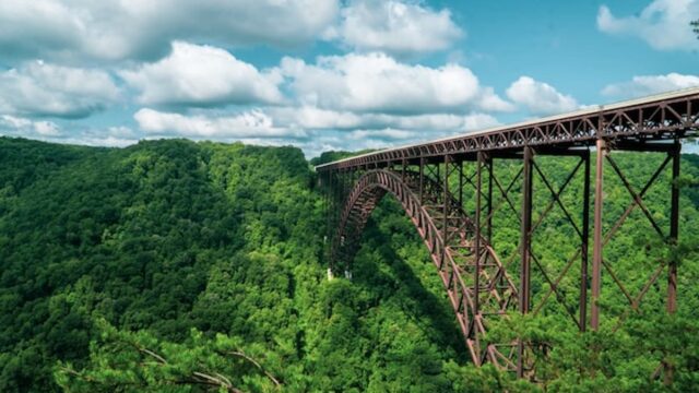 Steel arch bridge spanning a forested gorge under a blue sky with clouds in West Virginia.