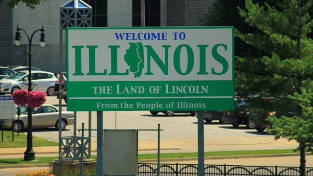 Green and white “Welcome to Illinois – The Land of Lincoln” roadside sign with trees and cars in the background