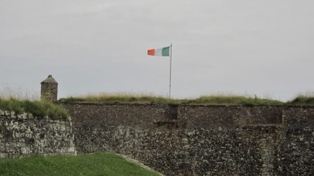 Irish tricolor flag flying above a stone fort wall under a cloudy sky in Ireland
