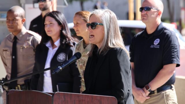 Arizona Governor Katie Hobbs speaking at a podium during an outdoor press conference with law enforcement officials standing behind her