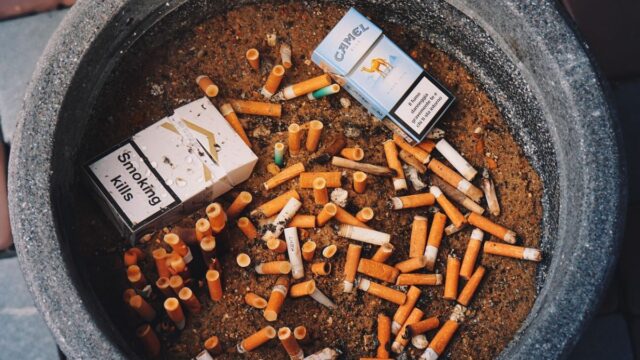 a top down close up of a large grey cement ashtray filled with sand, cigarette butts, and empty smoke packs