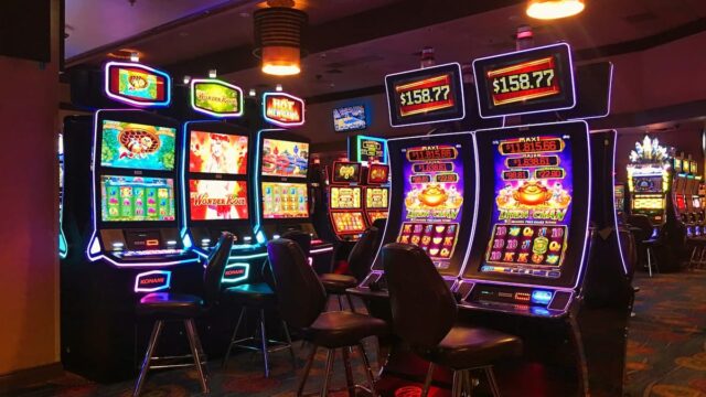 Row of illuminated slot machines on a casino floor with colorful screens and empty chairs in front