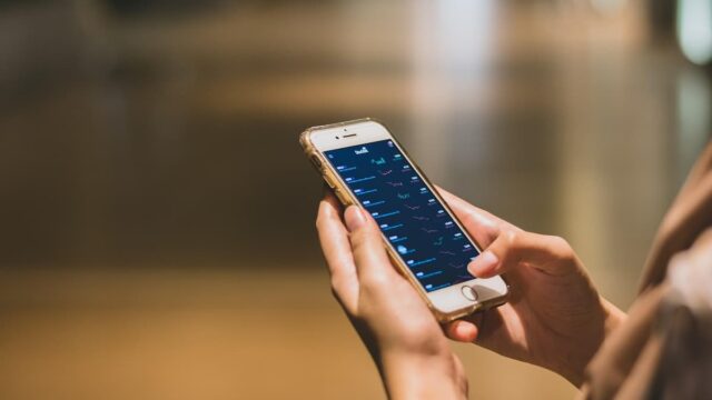 Person holding a smartphone displaying a financial trading app with stock price charts and market data on the screen.