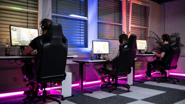 Three people sitting at desktop computers wearing headsets and playing video games in a neon-lit gaming room
