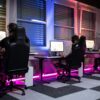 Three people sitting at desktop computers wearing headsets and playing video games in a neon-lit gaming room