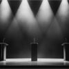 Three empty debate podiums under spotlights on a dark stage, representing US election politics
