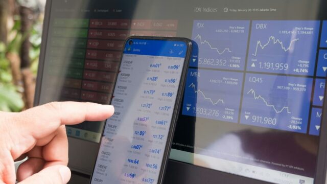Smartphone displaying financial trading quotes in front of a digital market dashboard, illustrating prediction market activity and contract settlement mechanics.