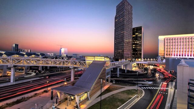 Aerial view of Tysons Corner Metro station at sunset, with red light trails from traffic, a pedestrian bridge, and office towers glowing against a pink and blue sky.