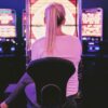 A blonde woman in a ponytail, white shirt, dark pants and sneakers sits back to the camera iin a chair in from of a slot machine with a line up of slot machines filling the background