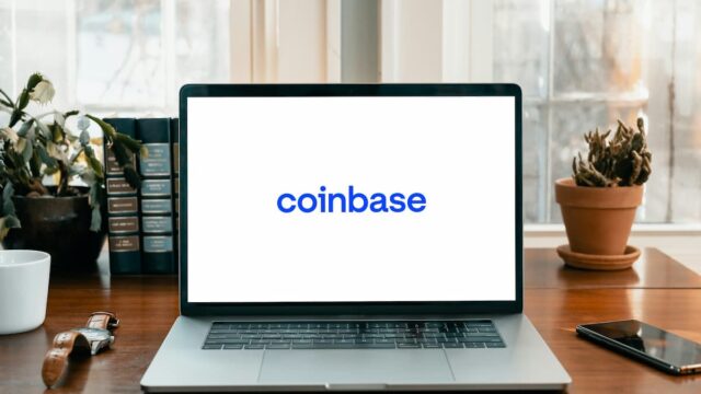 A laptop displaying the Coinbase logo on a desk beside a smartphone, books, plants, and a coffee cup.