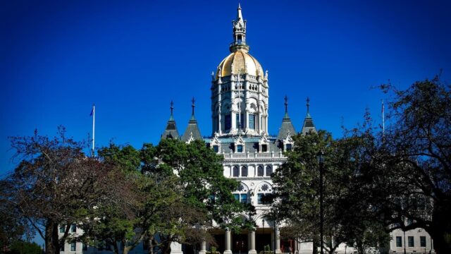 The Connecticut State Capitol building in Hartford under a clear blue sky with trees and a walkway in the foreground.