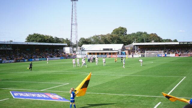 Players on a lower-league football pitch during a match at a small UK stadium with betting sponsor signage visible around the ground