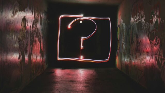 A light exposure drawing of a questiuon mark inside a square is visible at the ned of a long, dark, grafitti filled alley or hall