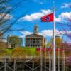 The Tennessee State Capitol building with Tennessee state flags and trees in the foreground under a blue sky.