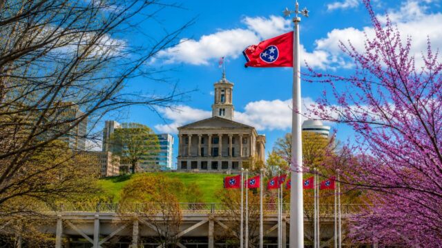 The Tennessee State Capitol building with Tennessee state flags and trees in the foreground under a blue sky.