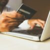 A person holding a payment card while using a laptop computer.