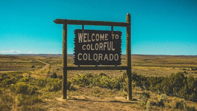 A large wooden sign against a bright blue sky on prairie-type land reads 'Welcome to Colorful Colorado'