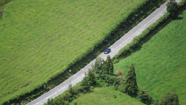 Lone black car travelling down a long asphalt road. On either side, road is banked by expansive green fields with border hedges