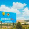 Welcome to New Mexico roadside sign with the state symbol and desert landscape under a blue sky.