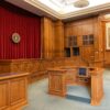 Interior of an empty courtroom with wooden benches, a judge's bench, and red curtains behind the dais.