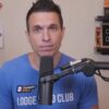 Doug Polk speaking into a microphone in a recording studio with poker memorabilia displayed on shelves behind him.