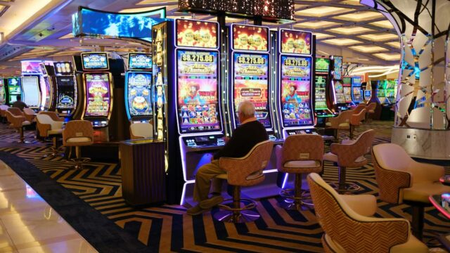 A patron plays slot machines inside a commercial casino gaming floor with rows of electronic gaming machines in the background.