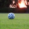 An NCAA-branded soccer ball resting on a grass field at sunset.