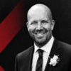 Josh Ercole of the CCGP smiles wearing a black suit with a white carnation in his lapel against a black background.