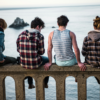 a group of mixed gender youth sit on a ledge overlooking a large body of water with backs to camera