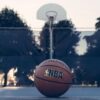 NBA basketball resting on an outdoor court with a hoop and trees blurred in the background.