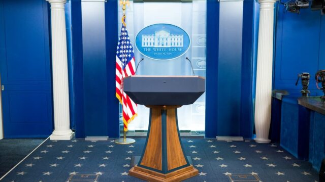 Empty podium in the White House press briefing room with a U.S. flag and television cameras.