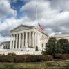 U.S. Supreme Court building under a cloudy sky with an American flag flying in front.