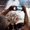 Fan holds up a smartphone to record a live soccer match in a crowded stadium.