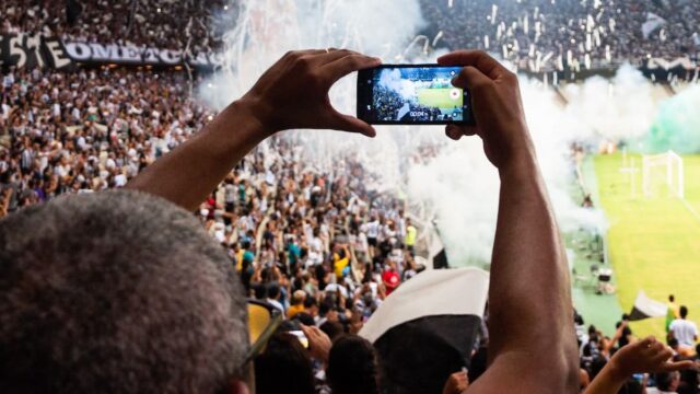 Fan holds up a smartphone to record a live soccer match in a crowded stadium.