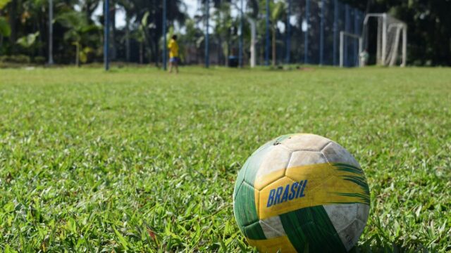 Brazil-themed soccer ball resting on a grassy soccer field with a goal and player in the background.