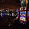 Rows of brightly lit slot machines on a casino floor with empty chairs in the foreground.
